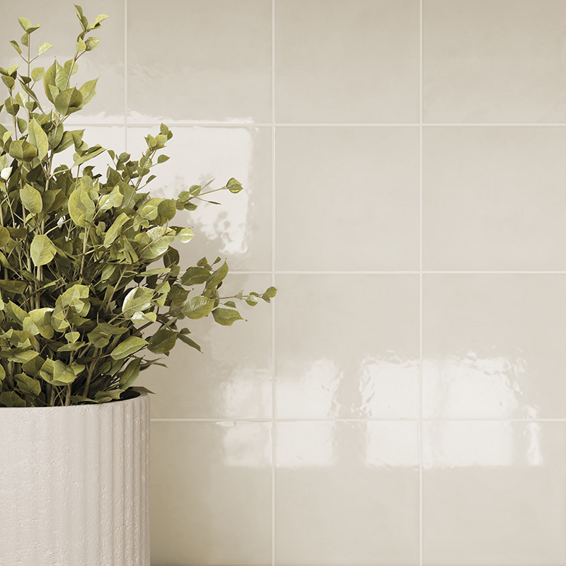 Azulejo cerâmico bege brilhante, com rejuntes formando quadrados; ao canto esquerdo, vaso cilíndrico creme com folhagem verde e reflexos suaves de luz sobre a superfície.