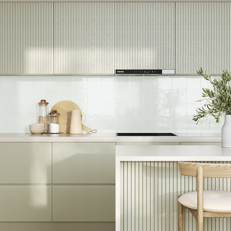 Carrelage mural blanc brillant à carreaux carrés; plan de travail clair, placards vert pâle à panneaux rainurés, accessoires de cuisine (bocaux, planche), vase à plante et tabouret bois.