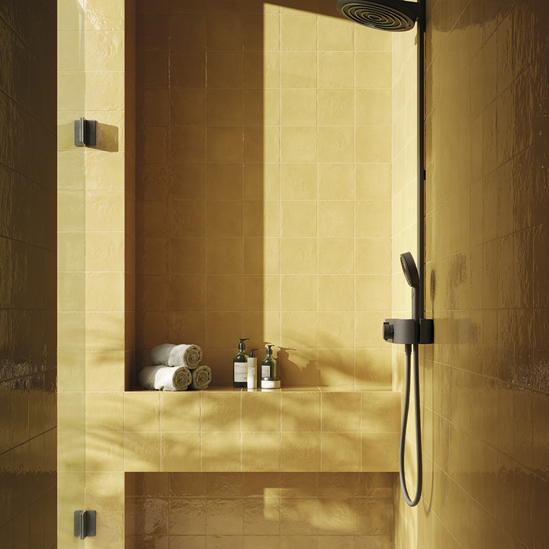 Azulejo amarelo em nicho de duche; bancada embutida com toalhas enroladas e frascos de sabonete, cabeçote de chuveiro de teto e ducha manual de parede.