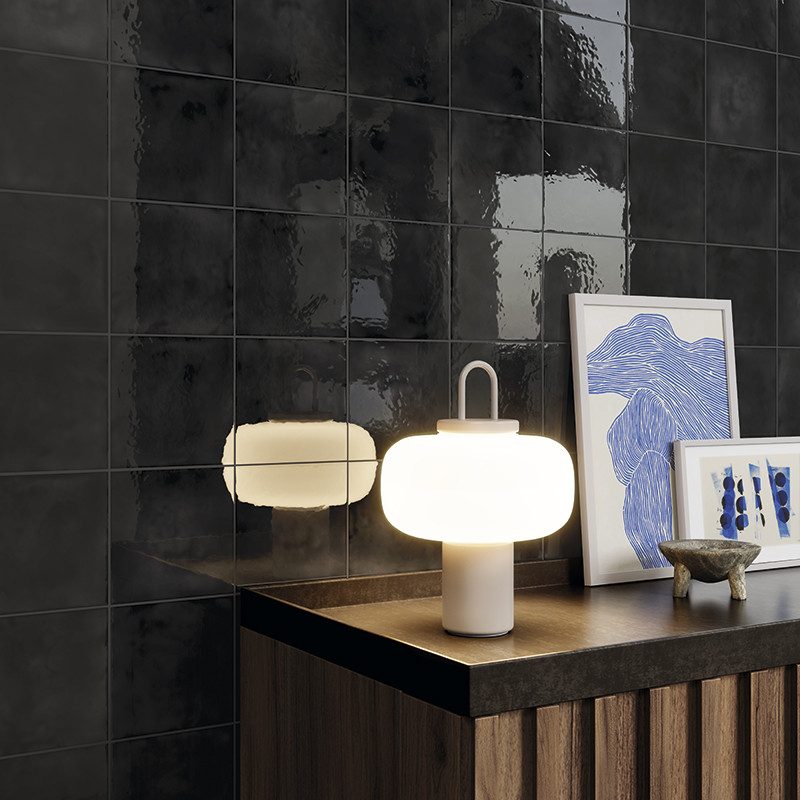 Glanzende zwarte vierkante wandtegels; context: houten dressoir met witte moderne tafellamp, reflectie in tegels, twee ingelijste blauw-witte abstracte prints en klein keramisch schaaltje.