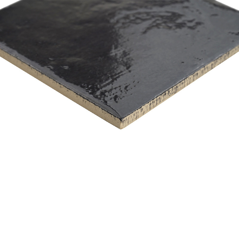 Glossy black glazed tile, unglazed beige edge visible; photographed in close-up on a white background, showing irregular texture and slight glaze shine.