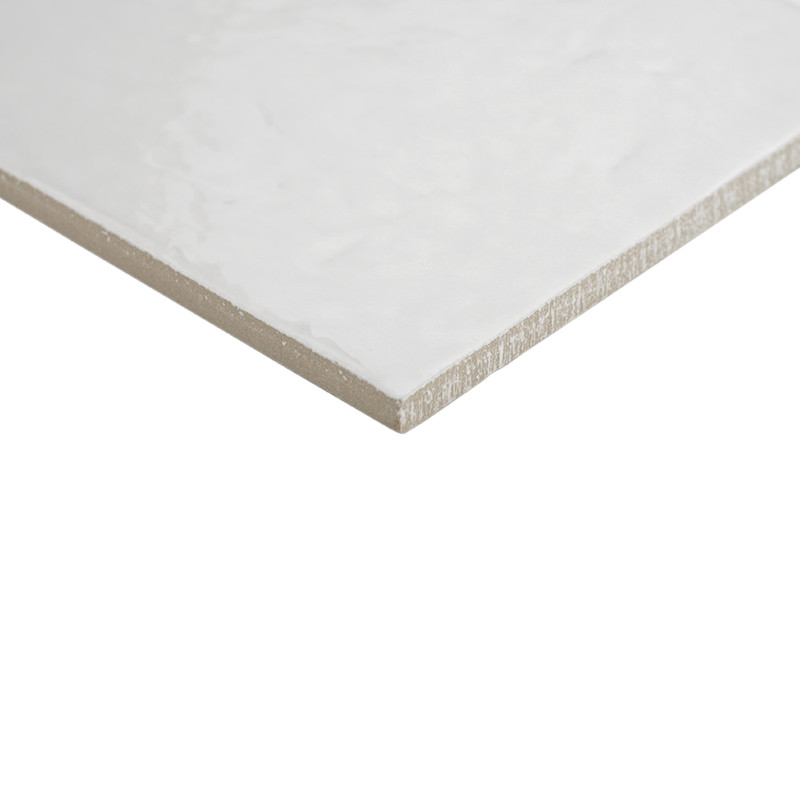 Azulejo branco brilhante com superfície sutilmente texturizada; borda exposta mostra corpo cerâmico; fotografado sobre fundo branco, enquadramento em perspectiva oblíqua.