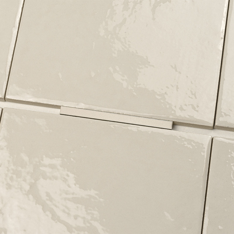 Beige glänzende Keramikfliese mit sichtbarer Fuge; kleiner rechteckiger Distanzhalter liegt in der Fuge; angrenzende gleiche Fliesen, leicht strukturierte, reflektierende Oberfläche.