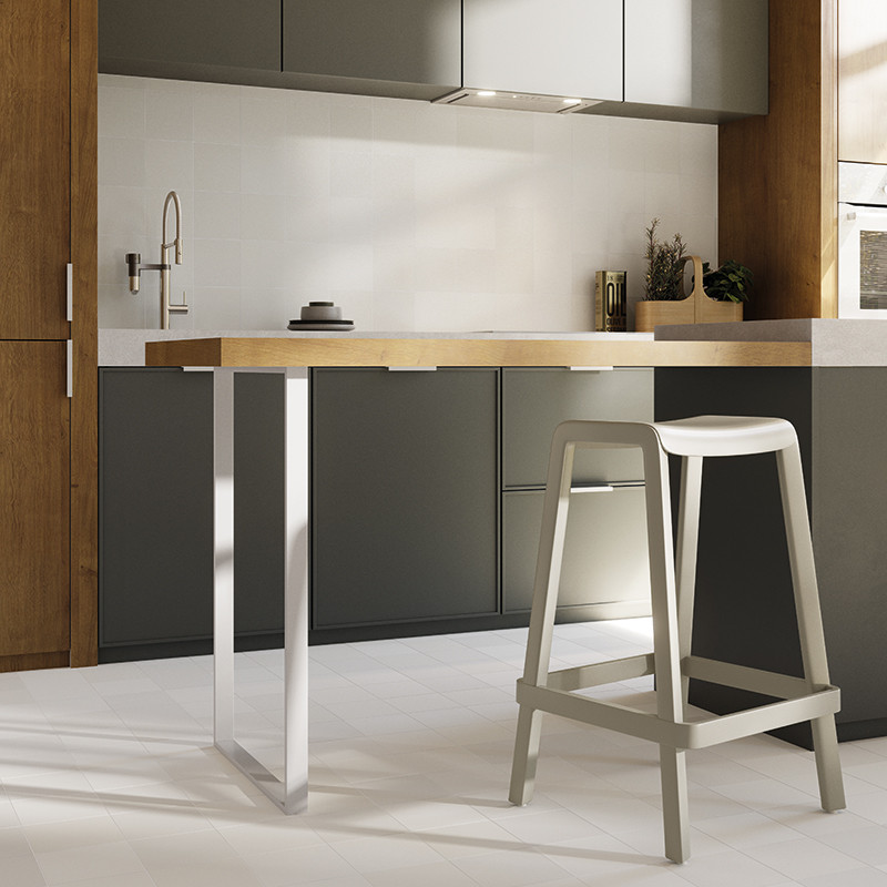 Carrelage gris clair grand format; cuisine moderne minimaliste avec îlot en bois à plateau clair soutenu par pieds métalliques, tabouret gris, meubles bas gris, robinet mural et crédence blanche.