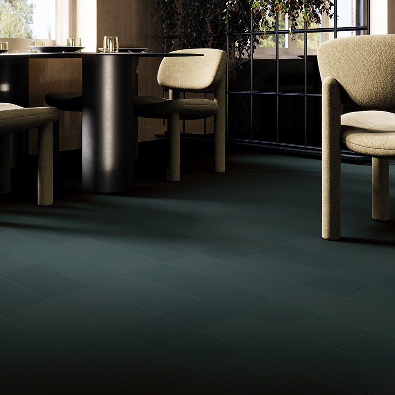 Dunkelgrüne, matte quadratische Bodenfliese mit feiner Karomusterung; Kontext: helles Café-Interieur mit runden schwarzen Tischen, gepolsterten Stühlen, großen Fenstern und Pflanzen.