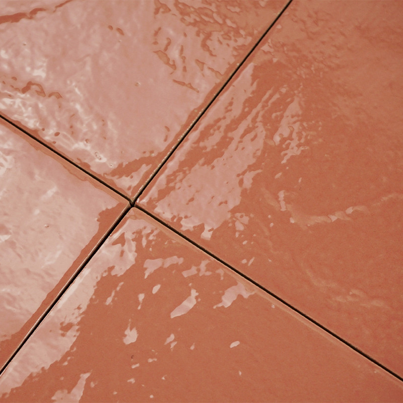 Azulejo esmaltado rojo ladrillo en primer plano; junta en cruz entre cuatro piezas, brillo reflectante con pequeñas irregularidades y manchas de humedad en la superficie.