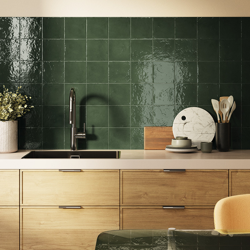 Carrelage mural vert foncé brillant (carrés) derrière un plan de travail de cuisine avec évier noir, robinet chromé, meubles en bois clair, ustensiles, plante et plateaux décoratifs.