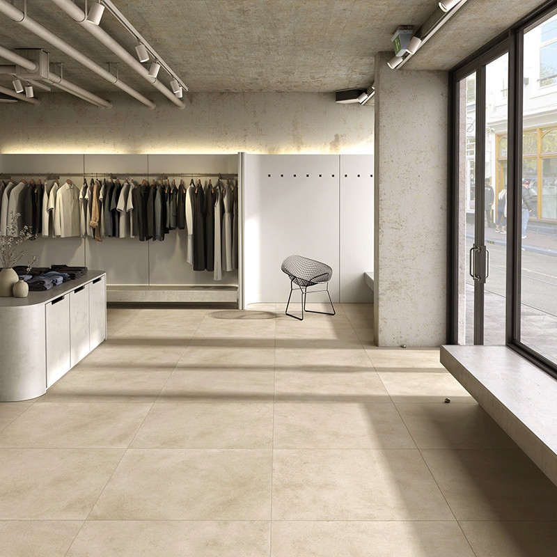 Baldosa cerámica beige mate en primer plano; tienda de ropa moderna con percheros, probadores blancos, silla metálica, escaparate de cristal y luz natural lateral.