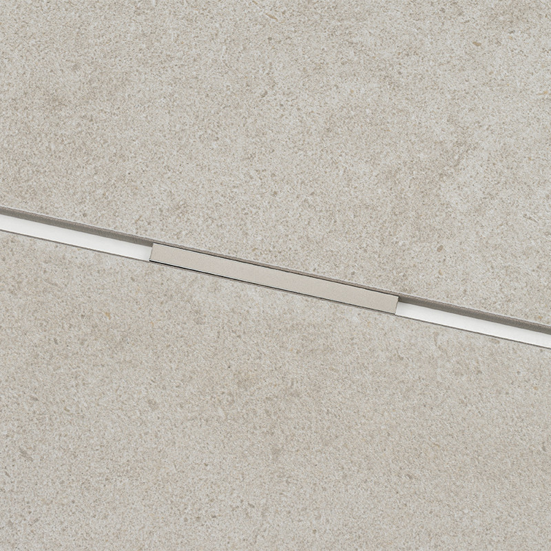Helle, beige, strukturierte Fliese mit diagonal verlaufendem, schmalem Metallprofil (Fuge/Abdeckung) mittig; körnige Beton- oder Keramikbodenoberfläche.