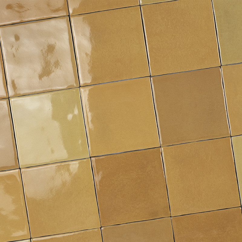 Azulejos cerámicos cuadrados color ocre brillante; dispuestos en filas con juntas oscuras, reflejos y sutiles variaciones tonales sobre una superficie plana.