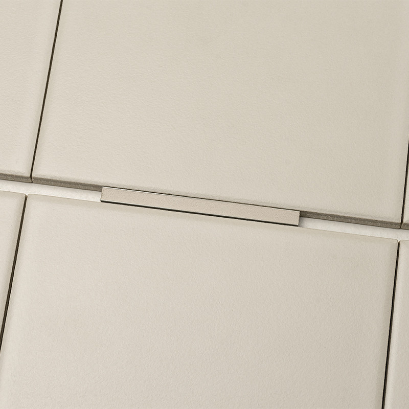 Carrelage beige mat : quatre carreaux carrés séparés par joints clairs, une plaquette rectangulaire métallique posée perpendiculairement sur un joint.