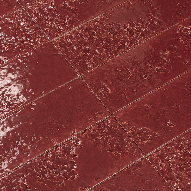 Carrelage rouge en dalles rectangulaires vernies au sol, joints visibles; surface irrégulière et rugueuse par endroits, brillante avec reflets.