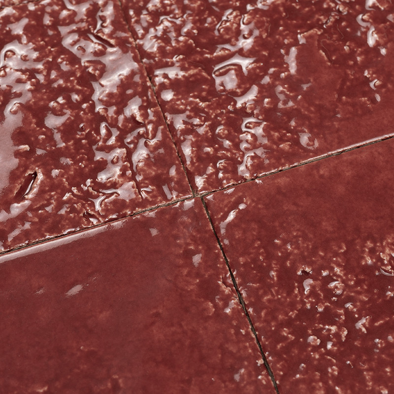 Carrelage rouge foncé à surface brillante et bosselée, joints fins formant une croix où quatre dalles se rejoignent, avec traces d'usure et reflets lumineux.