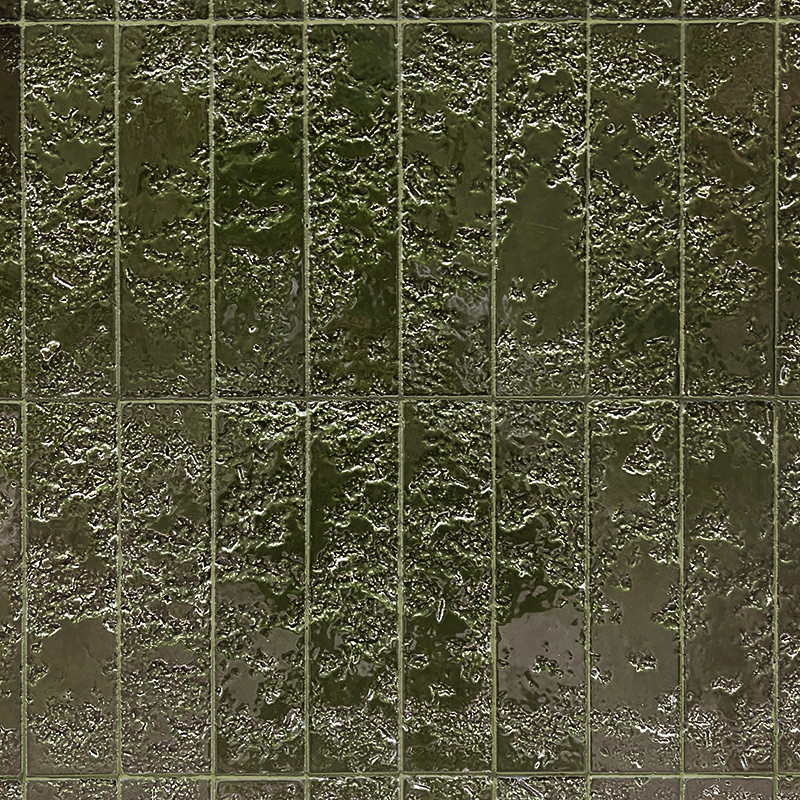 Azulejo vidriado verde oscuro con textura irregular y brillo húmedo; dispuesto en cuadrícula de finas juntas, sobre superficie vertical que sugiere pared interior.