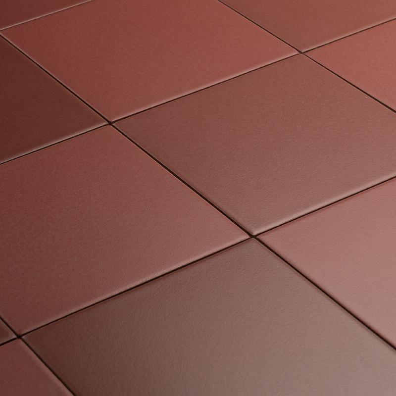 Carrelage quadrillé rouge-brun mat: grands carreaux carrés alignés, joints fins, surface légèrement texturée, perspective oblique montrant plusieurs rangées se rapprochant vers le fond.