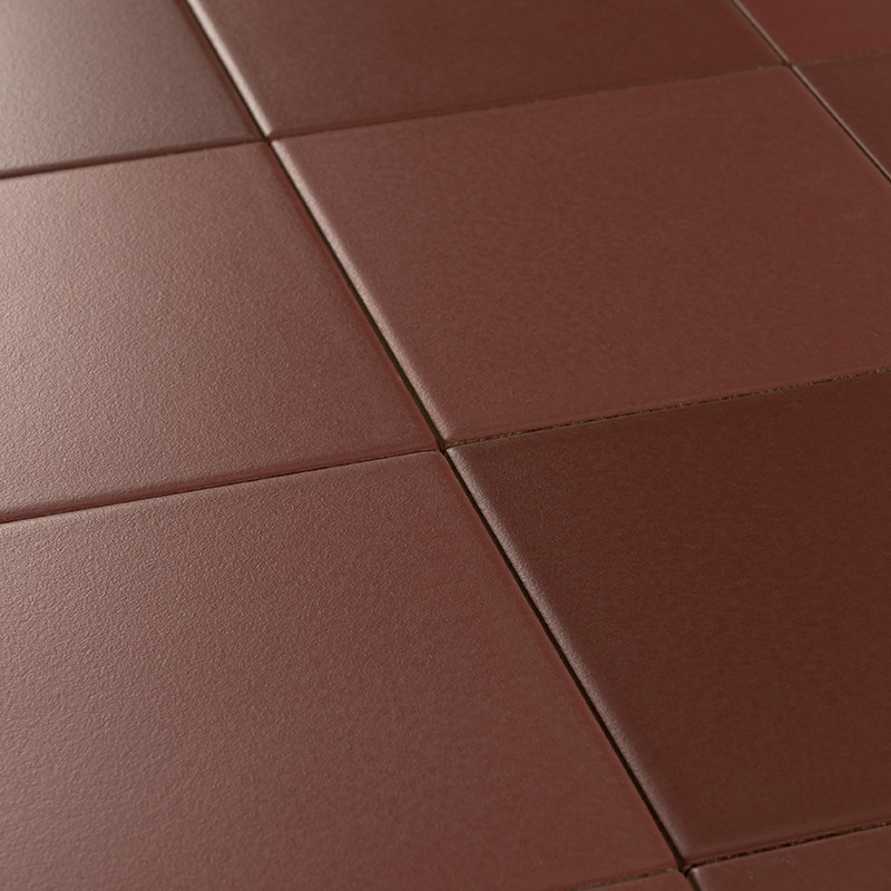 Carrelage de grandes dalles carrées brun rougeâtre, joints fins, surface mate légèrement texturée, perspective oblique montrant plusieurs carreaux se prolongeant vers l'arrière.