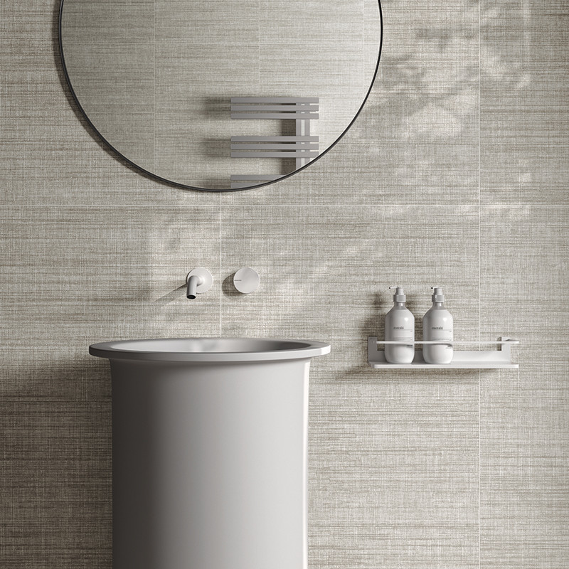 Azulejo texturizado beige: pared sobre lavabo pedestal gris, espejo circular, grifo empotrado y repisa con dos dispensadores; sombras suaves.