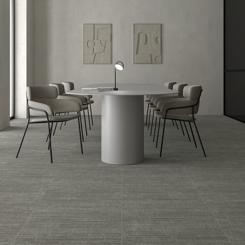 Azulejo de suelo gris texturizado en patrón cuadriculado; entorno: sala minimalista con mesa redonda cilíndrica central, ocho sillas tapizadas, lámpara de sobremesa y dos paneles murales abstractos.