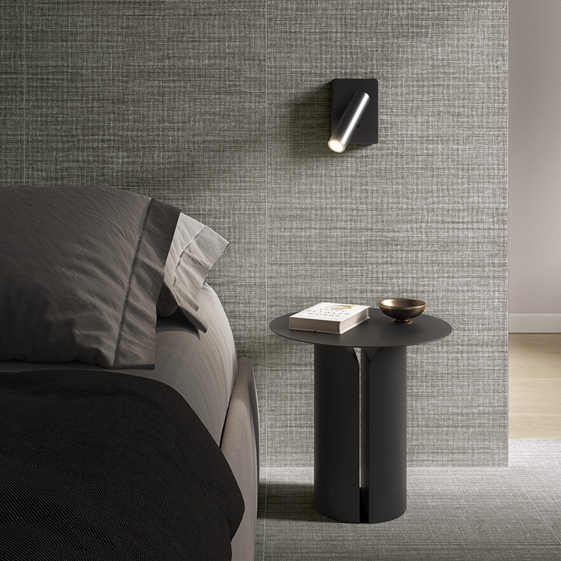 Azulejo texturizado gris en pared: junto a una cama oscura, mesita cilíndrica negra con libro y cuenco metálico; aplique mural negro inclinado y suelo de madera clara.