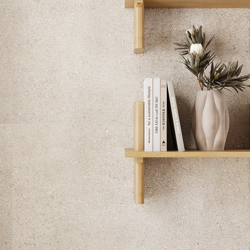 Carrelage mural beige à texture fine; étagères en bois sur la droite supportant trois livres et un vase beige sculpté contenant branches et fleurs séchées.