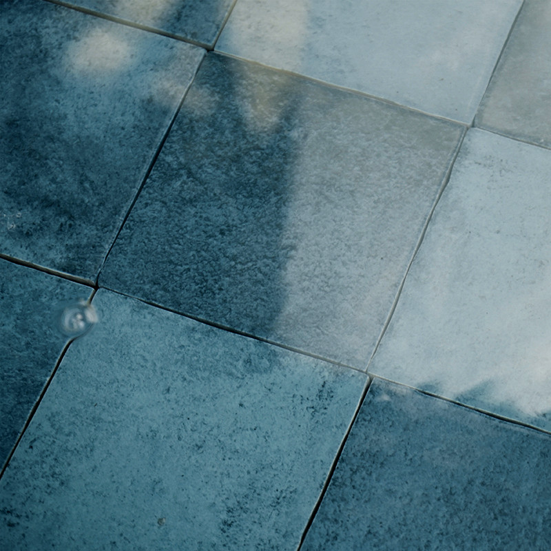 Carrelage de grandes dalles carrées bleu-gris; joints visibles, surface mouillée réfléchissante montrant ombre diffuse et une goutte créant petit anneau d'ondulation.