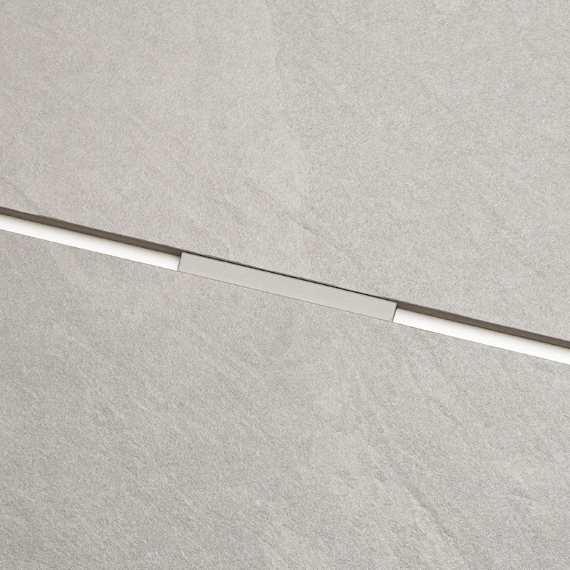 Light gray rectangular tile with stone texture; a narrow metal joint crosses diagonally, surroundings: continuous, matte, concrete-like surface.