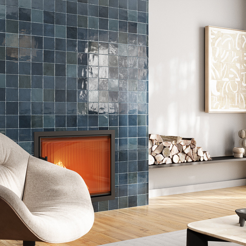 Carrelage bleu brillant encadrant une cheminée encastrée allumée; contexte: fauteuil beige près d'un sol en bois, rangée de bûches sur étagère et cadre mural clair.
