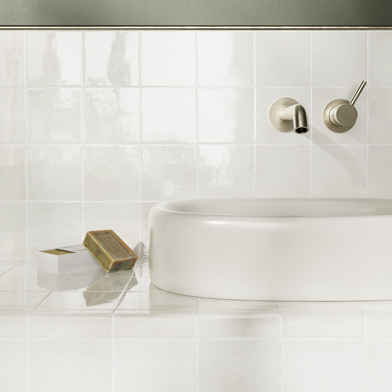Azulejos blancos cerámicos brillantes (pared y encimera); contexto: lavabo redondo sobre encimera, grifos metálicos empotrados y pastilla de jabón sobre la superficie.