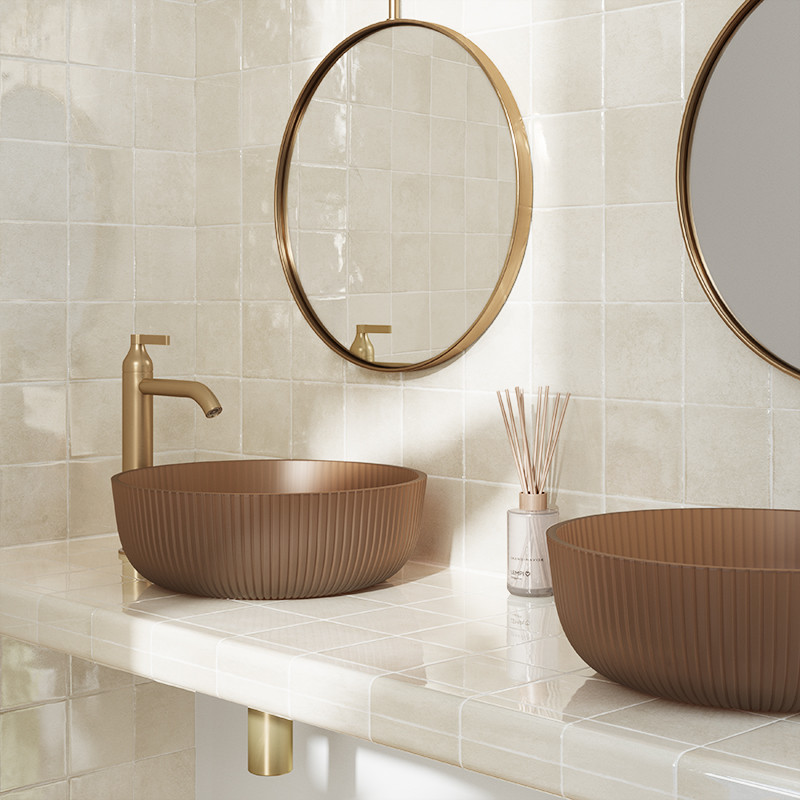 Quadratische beige Fliesen an Wand und Arbeitsfläche; braune gerippte Aufsatzwaschbecken, goldene Armaturen, runde goldgerahmte Spiegel und Duftstäbchen.
