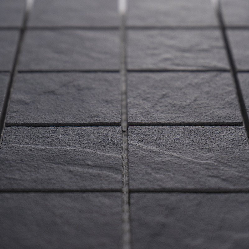 Azulejo cerámico oscuro con superficie ligeramente texturada y juntas claras, disposición en cuadrícula; entorno: más azulejos similares formando un suelo o pared.