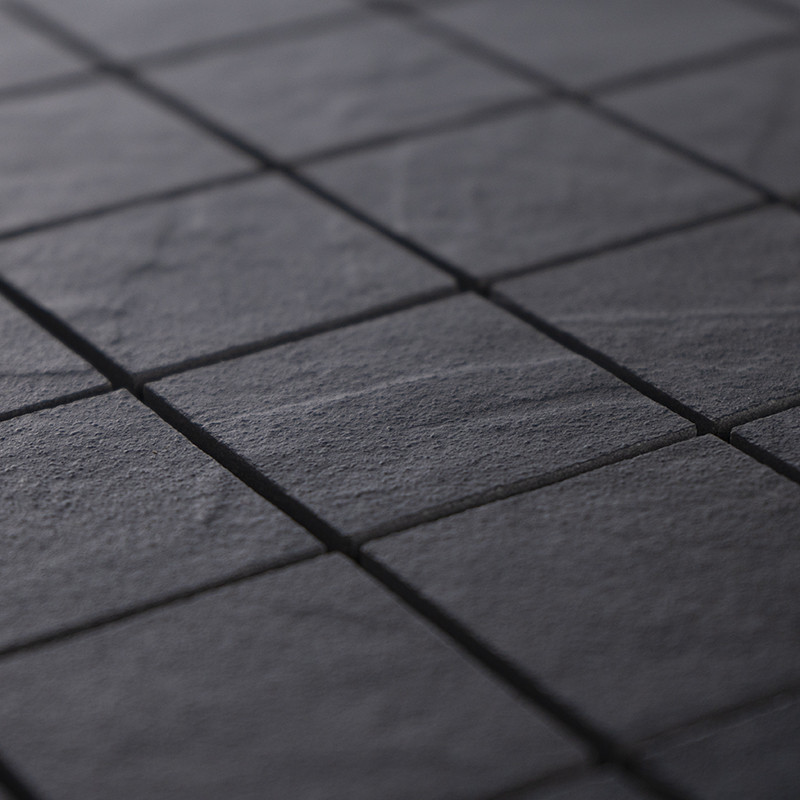 Square matte grey tile with a rough texture; dark joints create a grid, soft side lighting accentuates the relief, creating a sober and shaded atmosphere that suggests a nearby floor or wall.