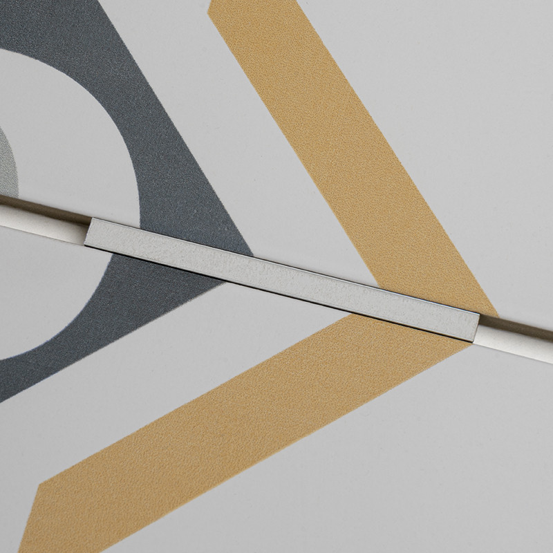 Fliese mit geometrischem Muster (graue Kreisform, goldene V‑Streifen) auf weißer Oberfläche; diagonale Fuge mit schmalem metallischem Abstandshalter über der Naht.