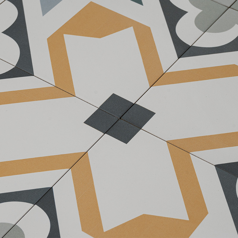 Fliesenfeld mit weißer Grundfläche und geometrischem Muster in Goldgelb und Grautönen; vier Fliesen treffen in einer dunklen, quadratischen Kreuzfuge auf einem Bodenbelag.