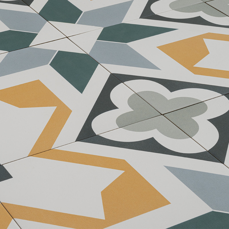Gemusterte Bodenfliesen mit geometrischen Stern- und Kleeblattmotiven in Grün, Grau und Senfgelb auf weißem Hintergrund; sichtbare Fugen, diagonal angeordnet.