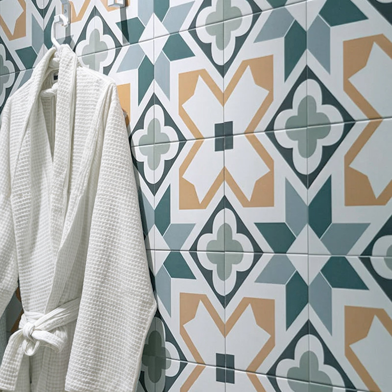 Geometrische Keramikfliesen in Grün, Beige und Weiß mit floral-kreuzförmigem Muster; links hängt ein weißer Waffelstoff-Bademantel an der Wand.