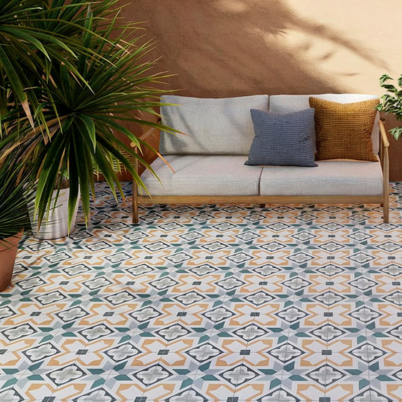 Gemusterte, farbige Bodenfliesen mit geometrischem Sternmuster (pastellblau, senfgelb, grau) vor hellem Sofa mit zwei Kissen; Schatten und große grüne Topfpflanzen am Rand.