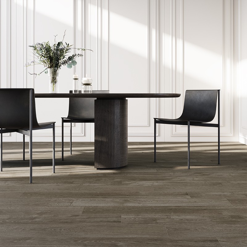 Carrelage imitation parquet brun-gris au premier plan; table ronde noire à base cannelée, trois chaises noires, vase de branchages et bougies, mur blanc à panneaux et lumière naturelle.