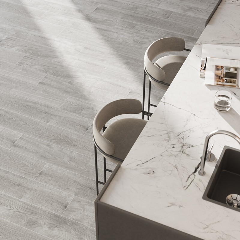 Encimera blanca de mármol con fregadero y grifo; contexto: suelo de lamas imitación madera claro, dos taburetes altos tapizados y luz natural diagonal.