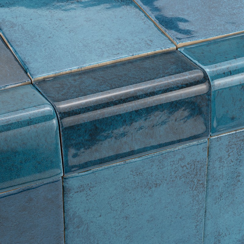 Glänzende blau-türkise Keramikfliese mit abgerundeter Eckkante und sichtbaren Fugen; dunkler, glänzender Stufenkantenstreifen durch Lichtreflexe.