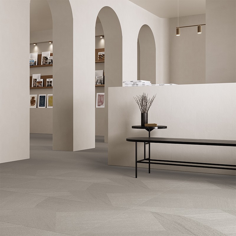 Baldosa beige mate en patrón geométrico; contexto: sala minimalista con arcadas blancas, banco negro con jarrón y estantes empotrados con cuadros.