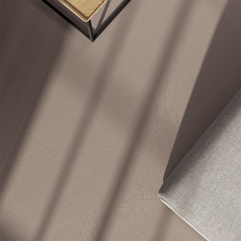 Azulejo rectangular beige con textura sutil; contexto: esquina de sofá gris claro a la derecha y sombras lineales de rejas proyectadas sobre el suelo.