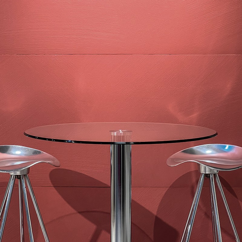 Table ronde en verre sur pied chromé, entourée de deux tabourets métalliques, devant un mur texturé couleur corail.