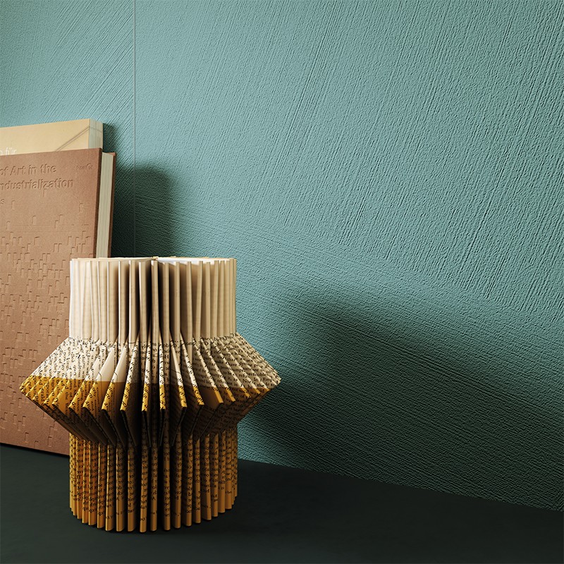 Sculpture of a folded book in the shape of a vase on a dark surface; turquoise textured wall in the background and a pale pink closed book leaning to the left.