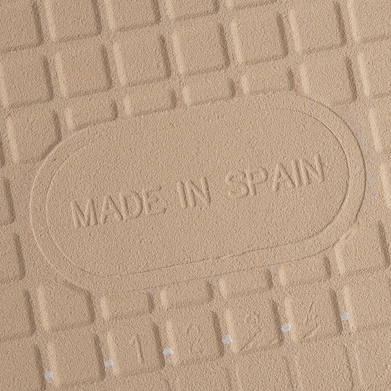 Carrelage beige texturé à motif quadrillé, inscription embossée « MADE IN SPAIN » centrée dans un ovale, petites perforations alignées en bas.