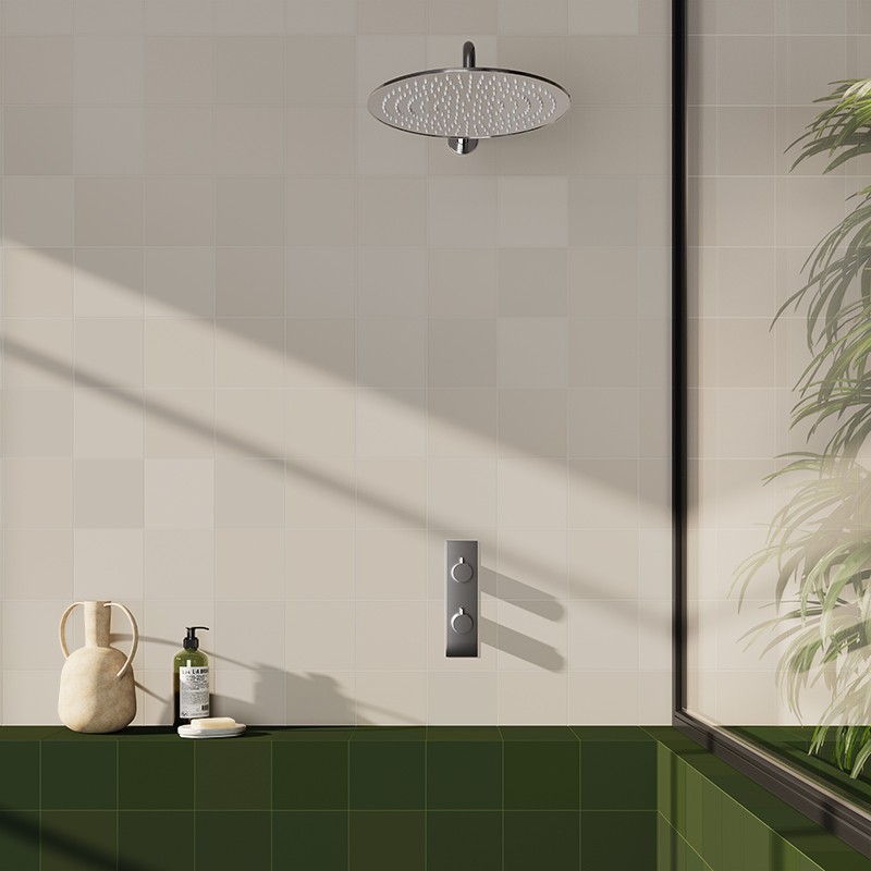 Azulejo cuadrado beige claro, en pared de ducha; ducha de techo cromada, placa de mandos, borde verde de bañera, frasco de jabón y planta junto a ventana con luz diagonal.