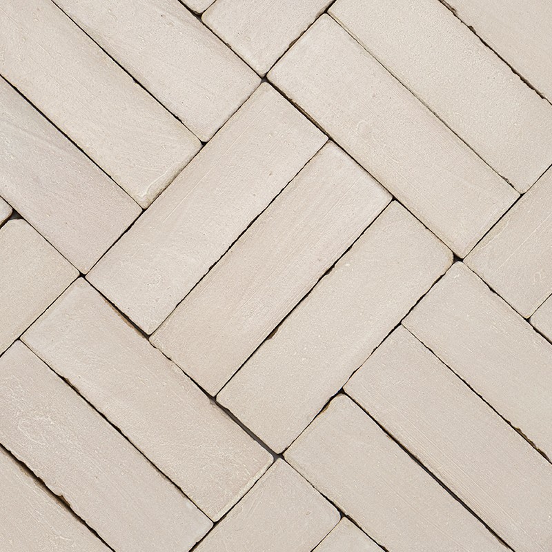 Azulejos retangulares bege-claro dispostos em padrão espinha de peixe; juntas finas e textura levemente irregular, sugerindo revestimento de parede ou piso interior.