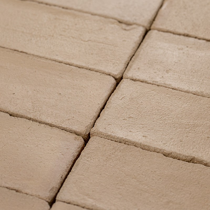 Azulejos rectangulares beige mate, con juntas profundas y bordes ligeramente irregulares; colocados en patrón de filas sobre superficie plana, con aspecto rústico y textura arenosa.