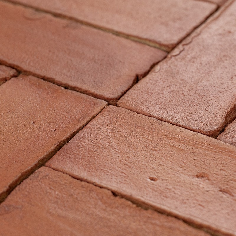 Terracotta-tegels met matte, licht korrelige oppervlakte, zichtbare voegen en kleine imperfecties; close-up van een vlakke vloer of wand, warm roodbruine kleur.