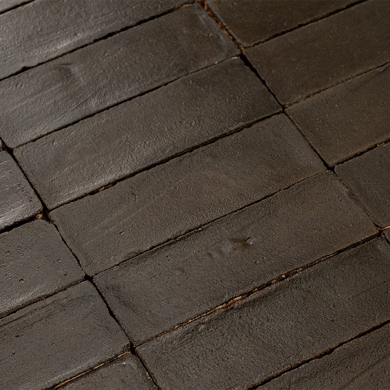 Dark rectangular tiles, matte surface with slight imperfections; arranged in regular rows with thin joints, aged ceramic appearance on interior floor or wall.