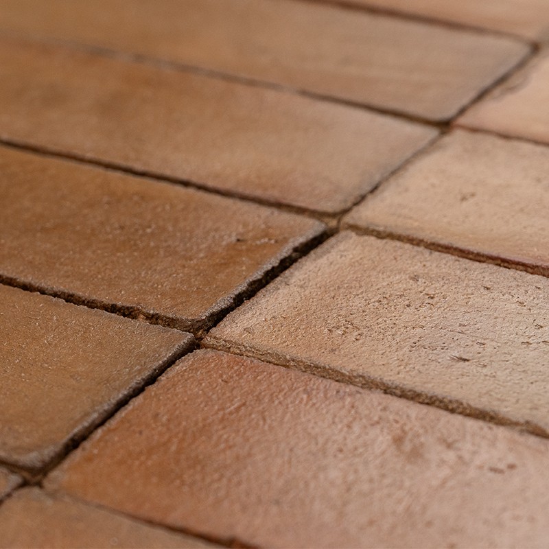 Azulejo rectangular de terracota, superficie mate y ligeramente desgastada; juntas oscuras entre piezas que forman un suelo rústico de tonos marrón rojizo.
